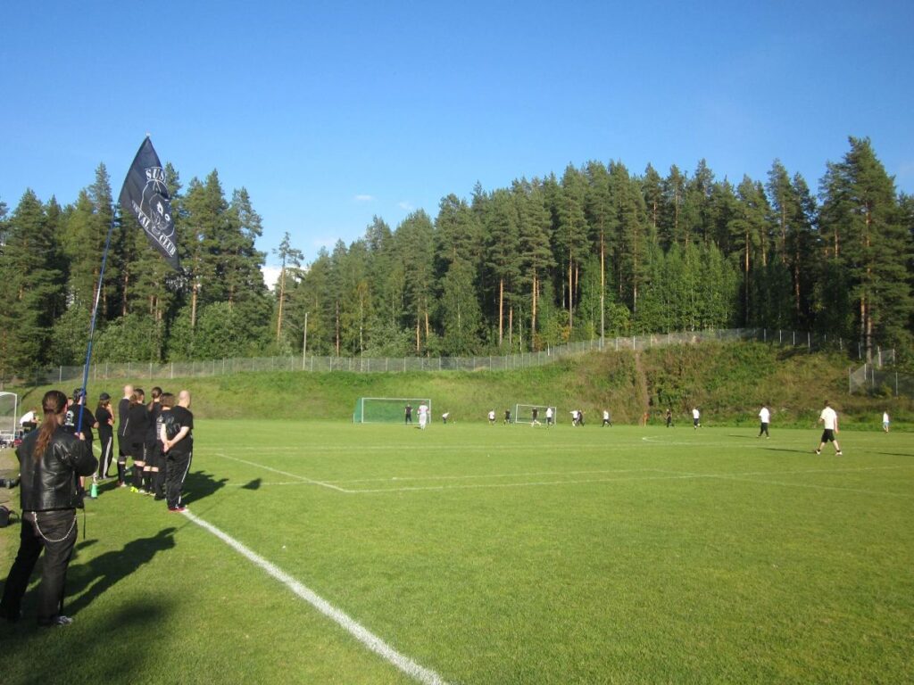 Jalkapallo-ottelu SMC vs. Ravintola La Barre kesällä 2014. SMC pelaa mustissa ja Barre valkoisissa. Etualalla myös SMC:n hallituksen puheenjohtaja SMC:n lipun kanssa.
