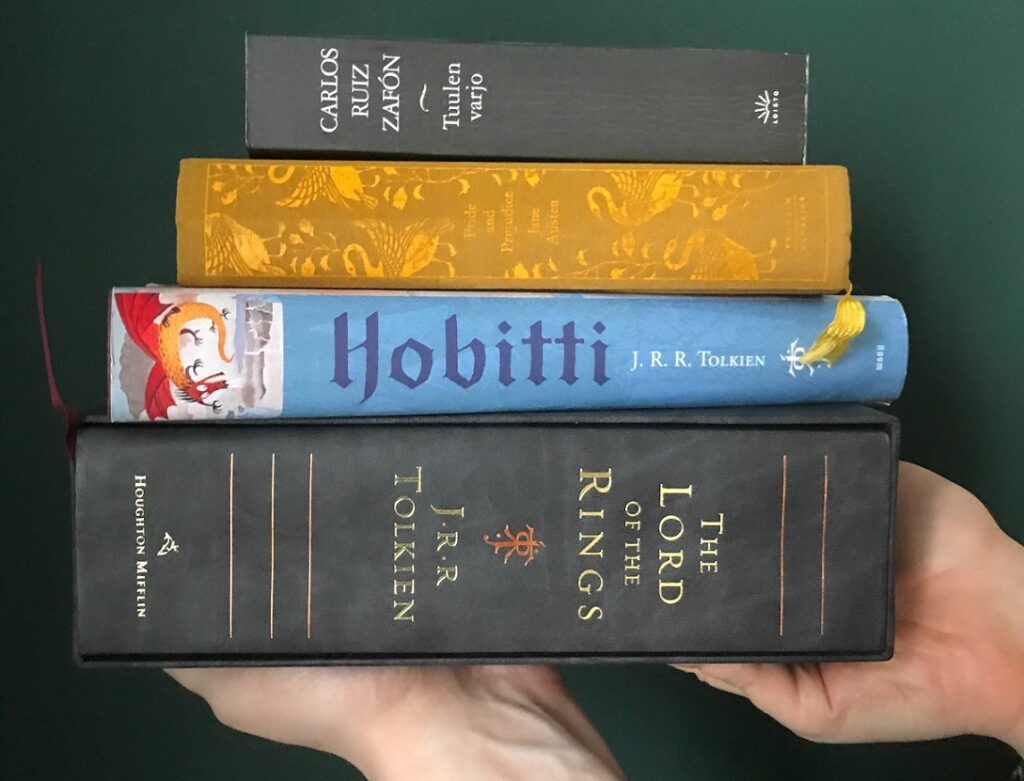 Kuvasssa vihreällä taustalla kädet jotka pitelevät neljän kirjan kirjapinoa. Kirjat ovat alhaalta ylöspäin The Lord of the Rings, Hobitti, Pride and Prejudice ja Tuulen varjo.